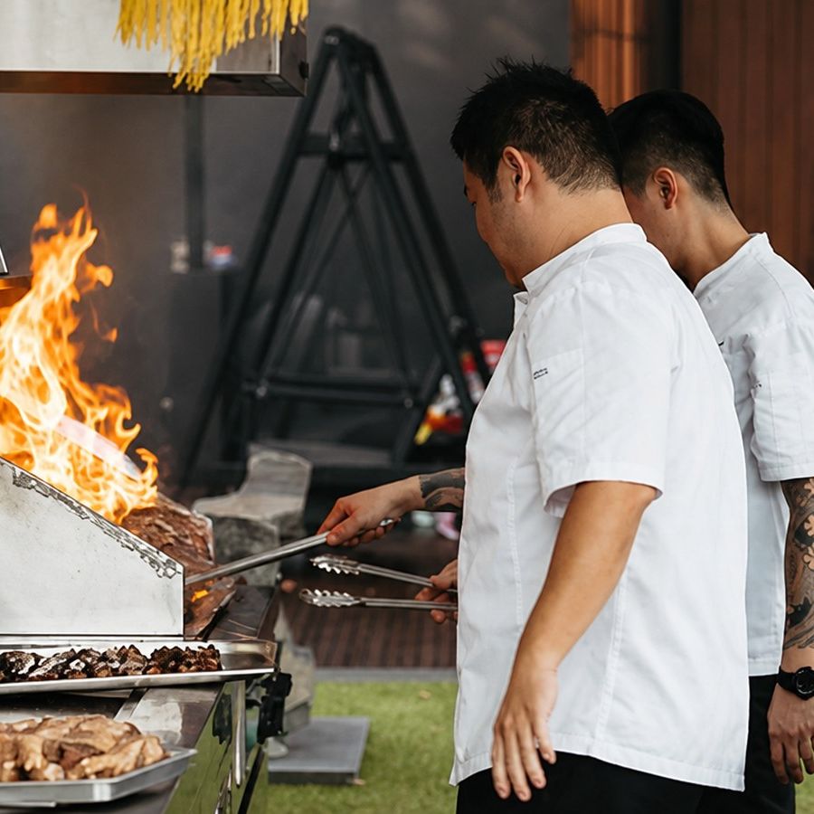 Two Shiok BBQ chefs grilling over an open flame at a live station in Singapore